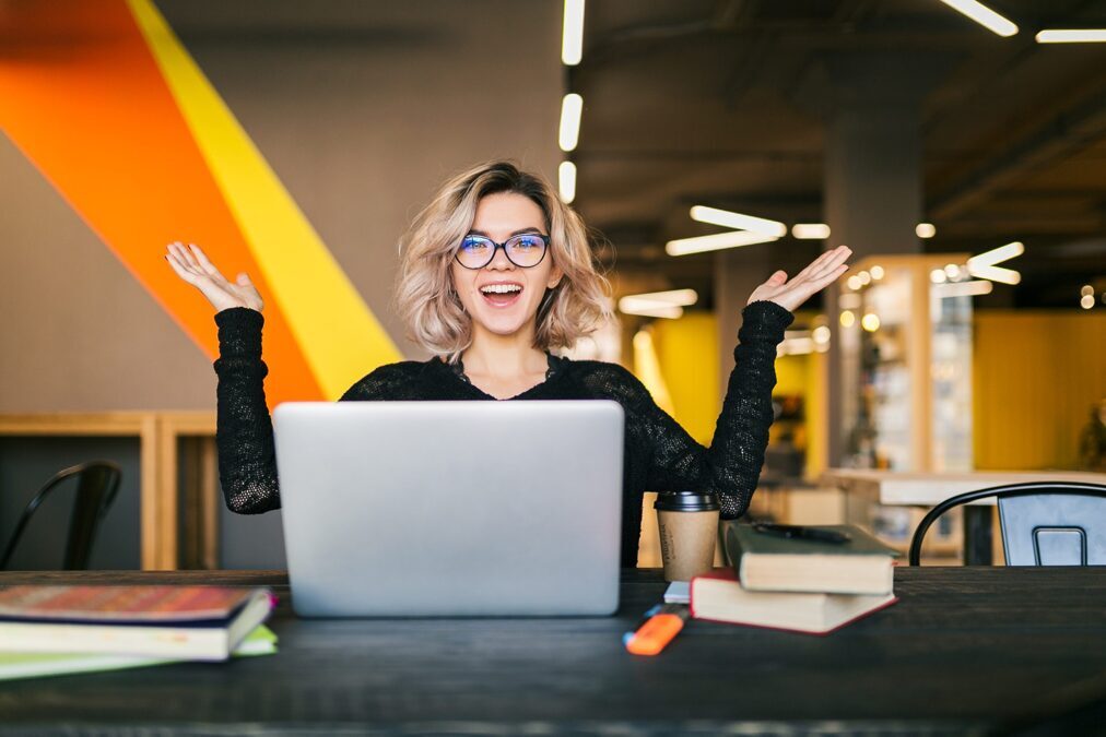 funny-happy-excited-young-pretty-woman-sitting-table-black-shirt-working-laptop-co-working-office-wearing-glasses-scaled